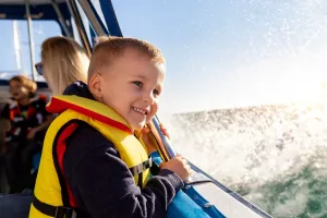 Portrait of cite little blond happy excited smiling caucasian boy wear lifevest enjoy sailing on motor boat sea against blue sky and water splash wave sun backlit. Summer travel vacation recreation.