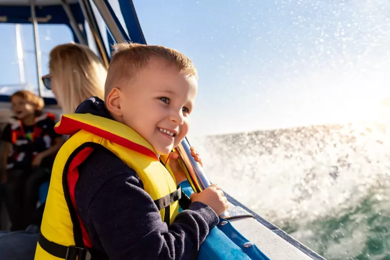 Portrait of cite little blond happy excited smiling caucasian boy wear lifevest enjoy sailing on motor boat sea against blue sky and water splash wave sun backlit. Summer travel vacation recreation.